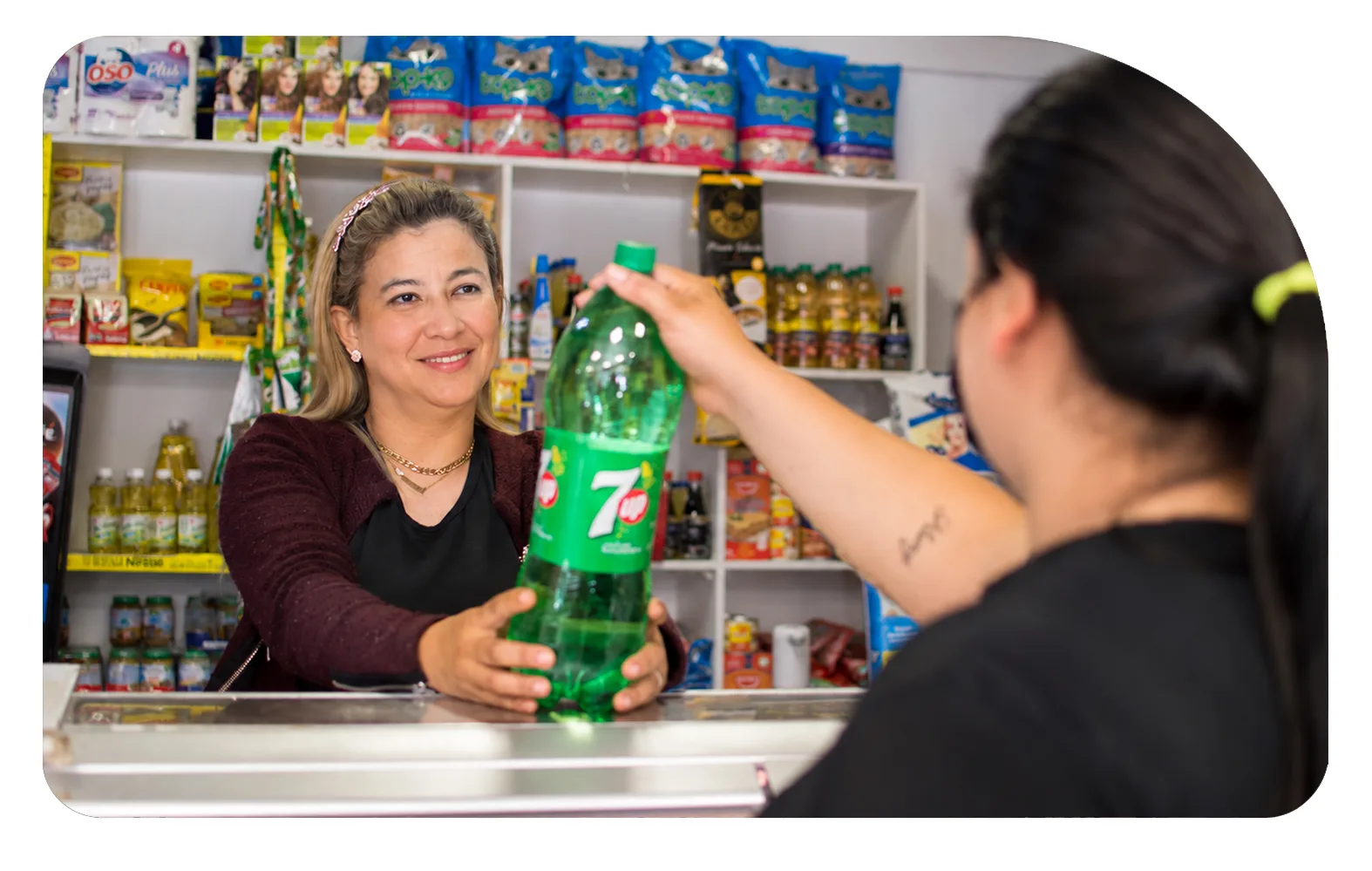 Almacenera entrega una botella de bebida a una clienta por sobre el mesón de su negocio